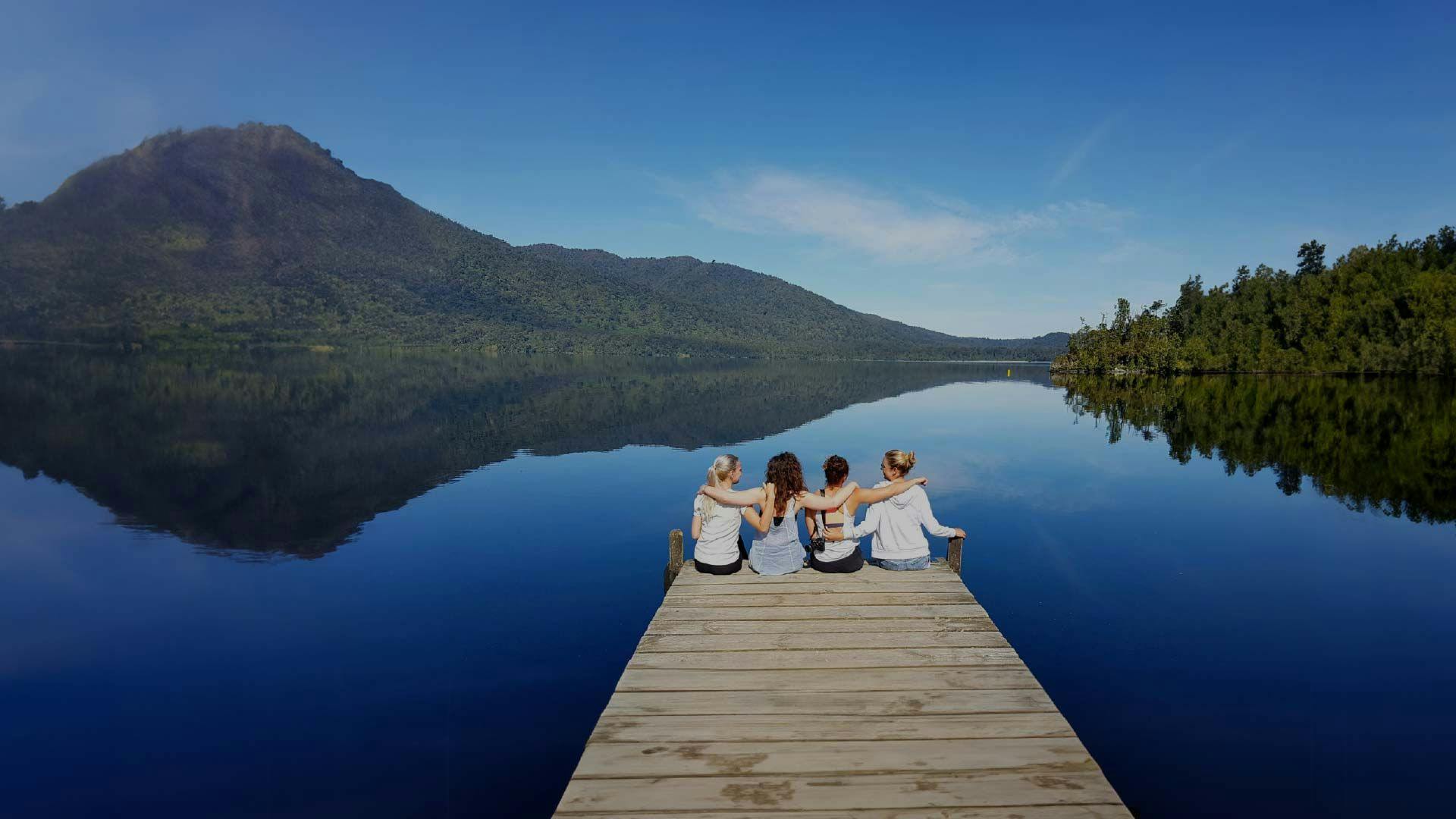 Four friends with their arms around in each other sat on a lake jetty in New Zealand