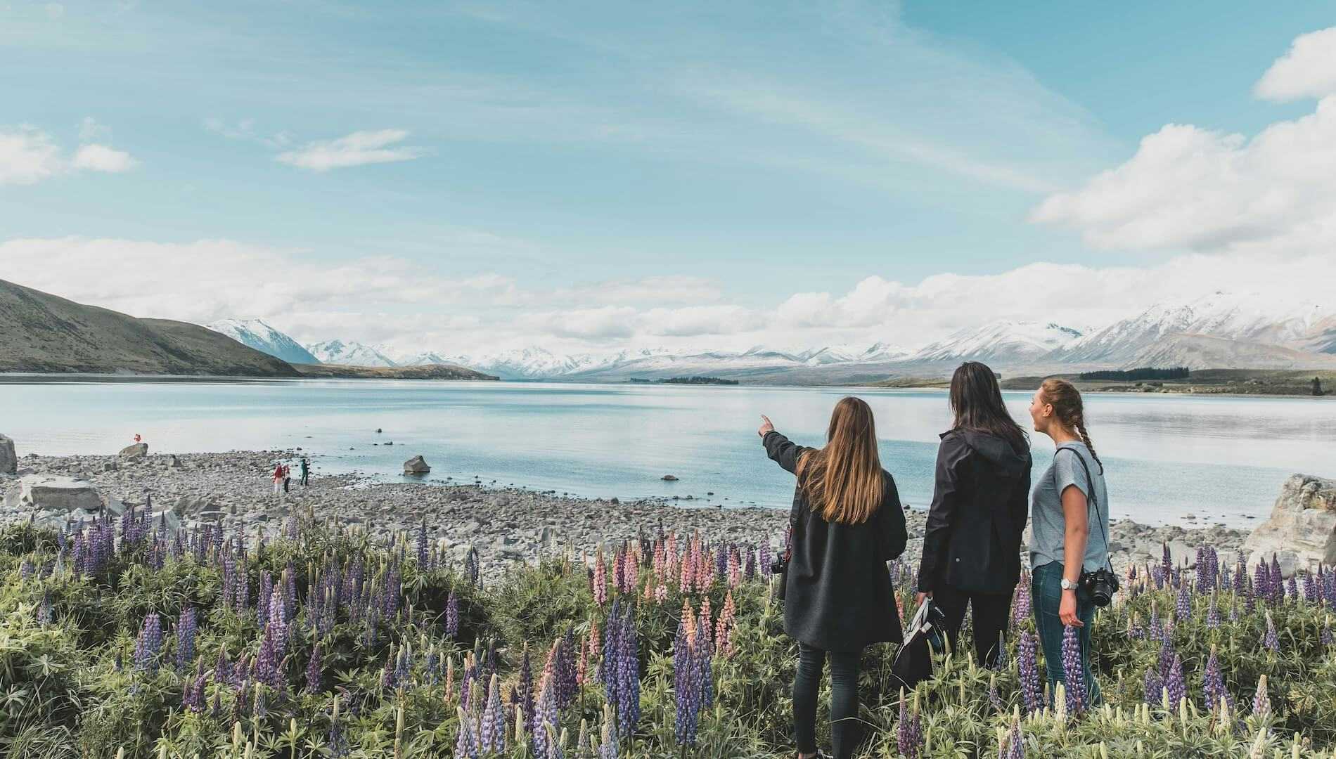 Three female travellers stand at Lake Tekapo looking out over snowy mountains surrounded by lupins during winter in New Zealand.