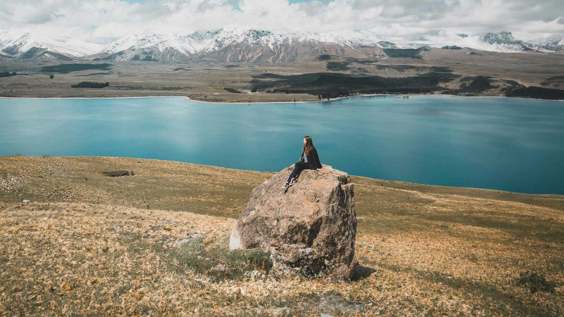 Woman sits on a large boulder admiring the view over Lake Tekapo